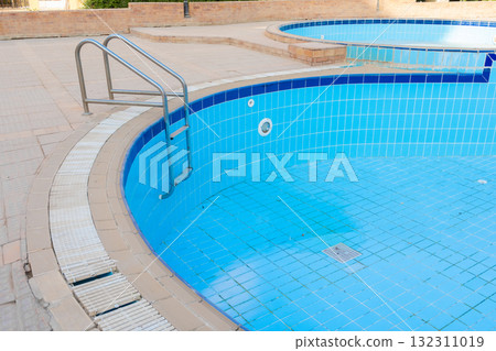 An empty swimming pool with blue tiles and a ladder. The water is absent, revealing the tiled bottom. Surrounding greenery is visible in the background. An empty swimming pool with blue tiles and a ladder. The water is absent, revealing the tiled bottom. Surrounding greenery is visible in the background. 132311019