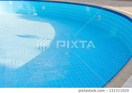 An empty swimming pool with blue tiles and a ladder. The water is absent, revealing the tiled bottom. Surrounding greenery is visible in the background. 132311020