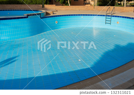 An empty swimming pool with blue tiles and a ladder. The water is absent, revealing the tiled bottom. Surrounding greenery is visible in the background. 132311021