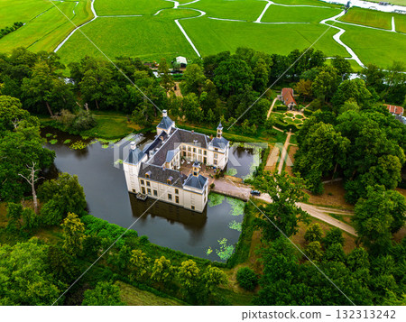 Aerial view of historic castle symmetrical twin towers and central hall framed by forest, moats, and tree lined path, with open fields and residential zone beyond under warm sunrise sky. Aerial view of historic castle symmetrical twin towers and central hall framed by forest, moats, and tree lined path, with open fields and residential zone beyond under warm sunrise sky. 132313242