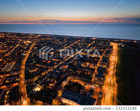 Aerial dusk view of coastal city streetlights and building lights glow against darkening sky, ocean meets horizon, structured grid of streets blends with natural sunset beauty. 132313248