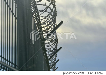 View of a prison fence topped with razor wire. Iron fence topped with barbed wire. Concept of guarded facilities, prisons, and military buildings. 132313262