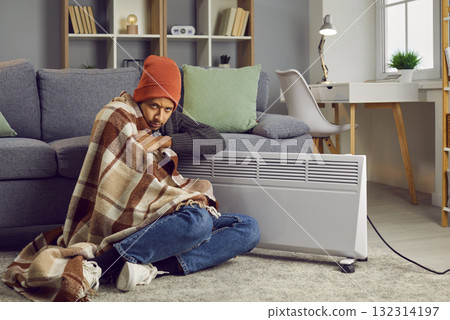 Disappointed man covered with blanket sitting on floor beside electric heater 132314197