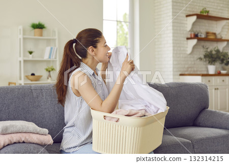 Happy young housewife sitting on sofa with laundry basket and smelling clean freshly washed clothes Happy young housewife sitting on sofa with laundry basket and smelling clean freshly washed clothes 132314215