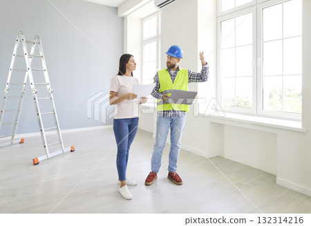 Builder in safety helmet talking to young woman in new house discussing renovation plans and repairs Builder in safety helmet talking to young woman in new house discussing renovation plans and repairs 132314216