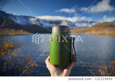 A green thermos for food in the hand of a tourist against the background of mountains A green thermos for food in the hand of a tourist against the background of mountains 132314850