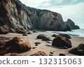 Rugged cliffs tower over a tranquil beach, where large rocks dot the sandy shore. Waves gently lap the coastline, creating a serene atmosphere at dusk 132315063
