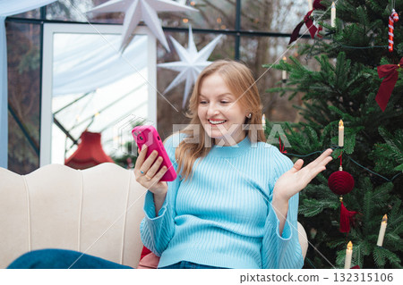 Happy blonde Caucasian woman having video call on smartphone near Christmas tree 132315106