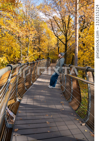 A woman in a gray jacket and beanie leans back on a modern wooden bridge surrounded by vivid yellow autumn trees in VDNH park A woman in a gray jacket and beanie leans back on a modern wooden bridge surrounded by vivid yellow autumn trees in VDNH park 132315224