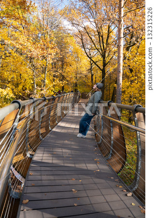 A woman in a gray jacket and beanie leans back on a modern wooden bridge surrounded by vivid yellow autumn trees in VDNH park A woman in a gray jacket and beanie leans back on a modern wooden bridge surrounded by vivid yellow autumn trees in VDNH park 132315226