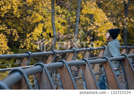 A woman in a green jacket and black hat stands on the ecological trail bridge in VDNH park surrounded by golden autumn leaves A woman in a green jacket and black hat stands on the ecological trail bridge in VDNH park surrounded by golden autumn leaves 132315234