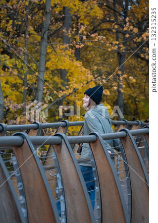 A woman in a green quilted jacket and black hat smiles while standing on a wooden bridge surrounded by yellow autumn foliage in VDNH park A woman in a green quilted jacket and black hat smiles while standing on a wooden bridge surrounded by yellow autumn foliage in VDNH park 132315235