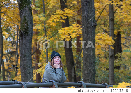 A woman in a green jacket and black hat rests on a wooden bridge railing, surrounded by golden maple leaves and tall trees in VDNH park A woman in a green jacket and black hat rests on a wooden bridge railing, surrounded by golden maple leaves and tall trees in VDNH park 132315248