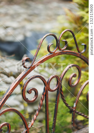Intricate red metal gate adorned with swirling designs opens to a lush garden surrounded by natural beauty 132315600