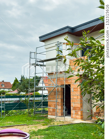 Workers on scaffolding repairing the exterior of a house surrounded by greenery on a cloudy day in a quiet neighborhood 132315616
