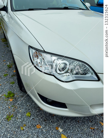 Shiny white car parked among pebbles, displaying bright headlights and sleek design under overcast sky 132315624