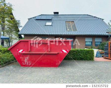Bright red construction dumpster stands out against a suburban house with gray shingles and a cloudy sky in early autumn 132315722
