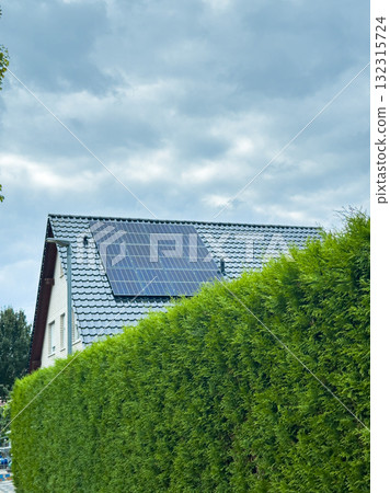 Bright green hedges frame a modern home showcasing solar panels under a cloudy sky in an eco-friendly neighborhood during the afternoon Bright green hedges frame a modern home showcasing solar panels under a cloudy sky in an eco-friendly neighborhood during the afternoon 132315724