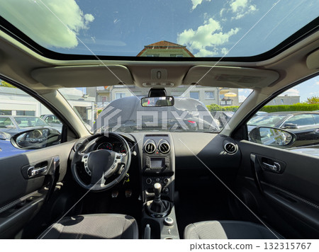 View from inside a parked car showing the dashboard and a glimpse of a dealership in bright daylight with clouds overhead 132315767