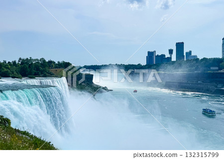 Majestic waters cascade over cliffs at Niagara Falls, with mist rising and boats gliding through the river, framed by a city skyline on a sunny day 132315799