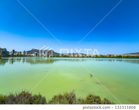 View of a serene lagoon with distant buildings and a bright blue sky in the background during a clear day in a coastal city View of a serene lagoon with distant buildings and a bright blue sky in the background during a clear day in a coastal city 132315808