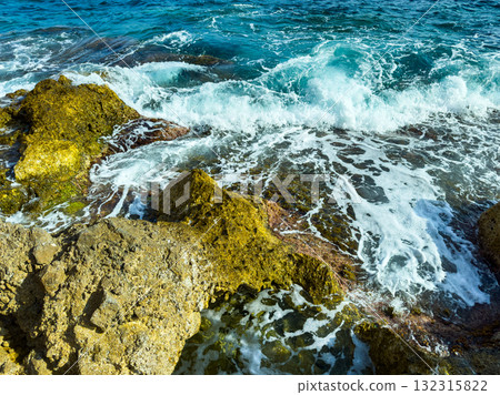 Waves crashing against rocky shore under bright blue sky with sparkling ocean during sunny day 132315822