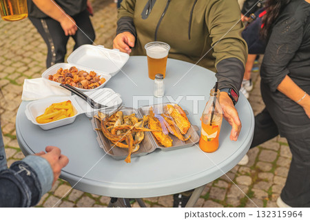 People enjoying fried fish, fries, and drinks at outdoor standing table during casual street meal. Concept of street food, fast food, holiday fair, disposable packaging, informal dining People enjoying fried fish, fries, and drinks at outdoor standing table during casual street meal. Concept of street food, fast food, holiday fair, disposable packaging, informal dining 132315964