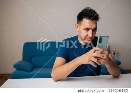 Smiling young man in casual clothes using mobile phone sitting at table, browsing internet or social media, concentrating on online content at home. Concept of modern communication, technology. 132316185