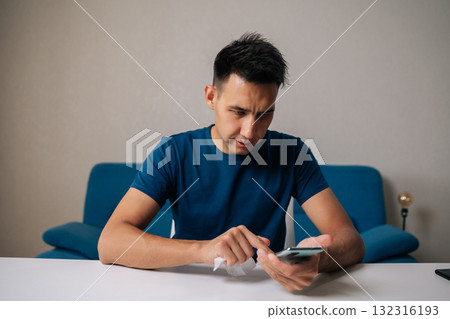Portrait of young man carefully cleaning smartphone screen with disinfectant wipe, maintaining hygiene and preventing spread of germs in home environment. Concept of healthy lifestyle. 132316193