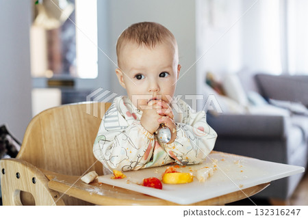 Cute baby learning to eat food with baby spoon by himself on high chair baby led weaning or blw. Infant self-feeding solid food, making mess, fine motor development. Kid enjoying healthy nutrition. Cute baby learning to eat food with baby spoon by himself on high chair baby led weaning or blw. Infant self-feeding solid food, making mess, fine motor development. Kid enjoying healthy nutrition. 132316247