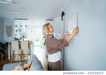 Side view middle aged woman putting painting picture in frame on the wall. Casual lady taking care of coziness in her new stylish apartment. Mature female decorating room. Moving and renovate house Side view middle aged woman putting painting picture in frame on the wall. Casual lady taking care of coziness in her new stylish apartment. Mature female decorating room. Moving and renovate house 132316253