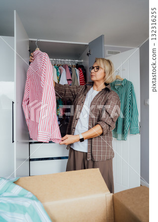 Middle aged woman selecting clothes from wardrobe for donating to Charity shop. Decluttering, Sorting clothes and Cleaning Up. Reuse, second-hand concept. Conscious consumer, sustainable lifestyle. Middle aged woman selecting clothes from wardrobe for donating to Charity shop. Decluttering, Sorting clothes and Cleaning Up. Reuse, second-hand concept. Conscious consumer, sustainable lifestyle. 132316255