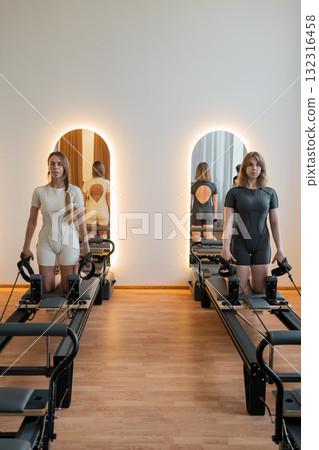 Split training of two girls adjusting reformer arm and core exercises, Woman performing Pilates exercises on reformer machine in gym Split training of two girls adjusting reformer arm and core exercises, Woman performing Pilates exercises on reformer machine in gym 132316458
