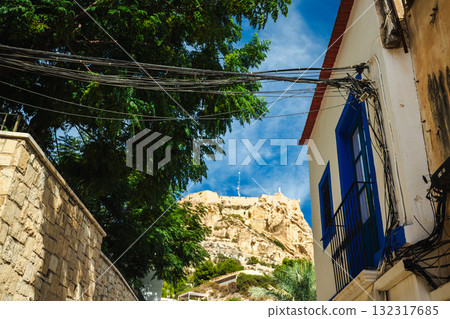 View of the Santa Barbara Castle above the traditional houses of the Santa Cruz district in Alicante, Spain, framed by a stone wall, green trees, and blue sky. 132317685