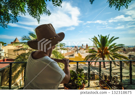 Woman in a hat enjoys the panoramic view of Alicante, Spain, from a shaded balcony surrounded by palm trees and potted plants on a sunny day. Woman in a hat enjoys the panoramic view of Alicante, Spain, from a shaded balcony surrounded by palm trees and potted plants on a sunny day. 132318675