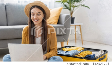 Young woman planning vacation using laptop with packed suitcase nearby 132321721