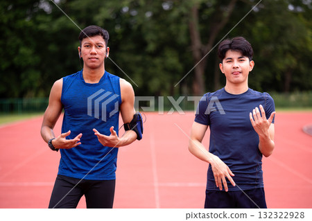 Two Asian men in sportswear standing on a running track, posing confidently before a workout session. Healthy lifestyle, teamwork, motivation, and outdoor fitness training for active individuals 132322928