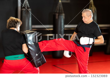 Elderly man practices kicking and punching with a trainer in gym 132323069