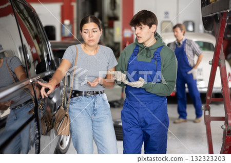 Upset girl owner of car points to scratches on car after an accident to an auto mechanic in car service center Upset girl owner of car points to scratches on car after an accident to an auto mechanic in car service center 132323203