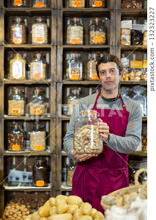 Grocery store employee in an apron offers nuts. Holds jar of nuts in his hands 132323227