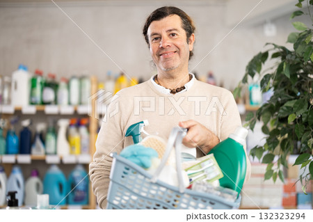 Positive man selects various household cleaning products for cleaning the house with full basket of various purchases 132323294
