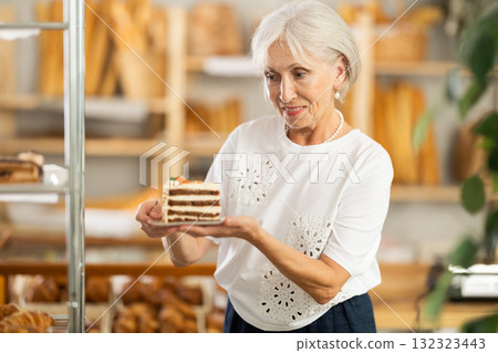 Senior female client takes piece of puff pastry cake from shelf of showcase, examines products Senior female client takes piece of puff pastry cake from shelf of showcase, examines products 132323443