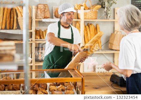 Man entrepreneur, owner of baker puts baguettes in paper bag, serves senior female client Man entrepreneur, owner of baker puts baguettes in paper bag, serves senior female client 132323454