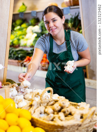 Woman seller wearing apron arranges garlic and other vegetables on counter and display case 132323462