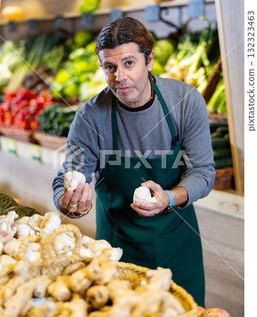 Man in supermarket seller in an apron carefully lays out fresh garlic on the shelves of store 132323463