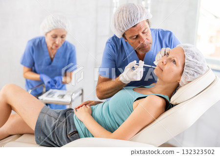 Male doctor with nurse help gives injection of botulinum toxin to client in lip area 132323502
