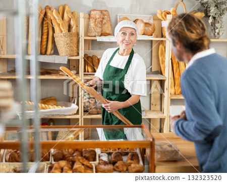 Mature woman entrepreneur, owner of baker puts baguettes in paper bag, serves male client Mature woman entrepreneur, owner of baker puts baguettes in paper bag, serves male client 132323520