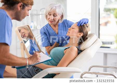 Elderly lady dermatologist examines skin of womans face, male nurse records data in client card 132323558