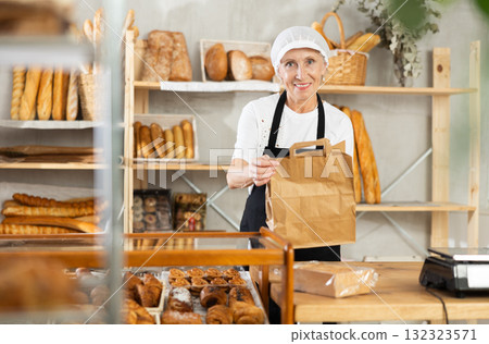Portrait of helpful female baker with paper bag of fresh baked goods in interior of bakery 132323571