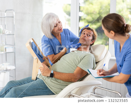Elderly lady dermatologist examines skin of mans face, female nurse records data in client card Elderly lady dermatologist examines skin of mans face, female nurse records data in client card 132323585